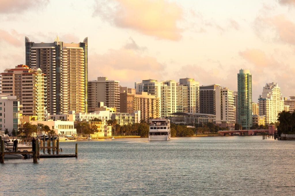 A boat sails through the canal in Miami Beach, with buildings silhouetted against the sunset