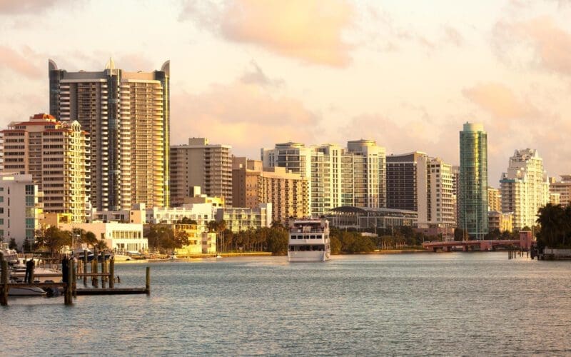 A boat sails through the canal in Miami Beach, with buildings silhouetted against the sunset