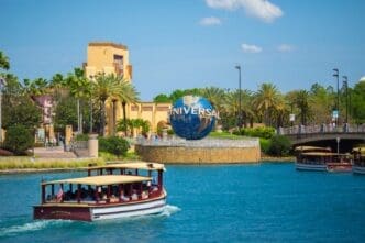 The Universal Studios globe in Orlando, Florida, with boats on the water and theme park buildings and palm trees in the background under a blue sky.