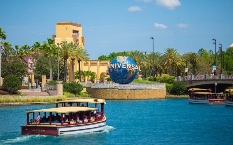 The Universal Studios globe in Orlando, Florida, with boats on the water and theme park buildings and palm trees in the background under a blue sky.