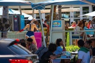 A wide shot of the Clevelander South Beach hotel and bar, with a woman in a sailor costume dancing. Crowds of people are seated at tables in a lively outdoor setting with umbrellas and palm trees.