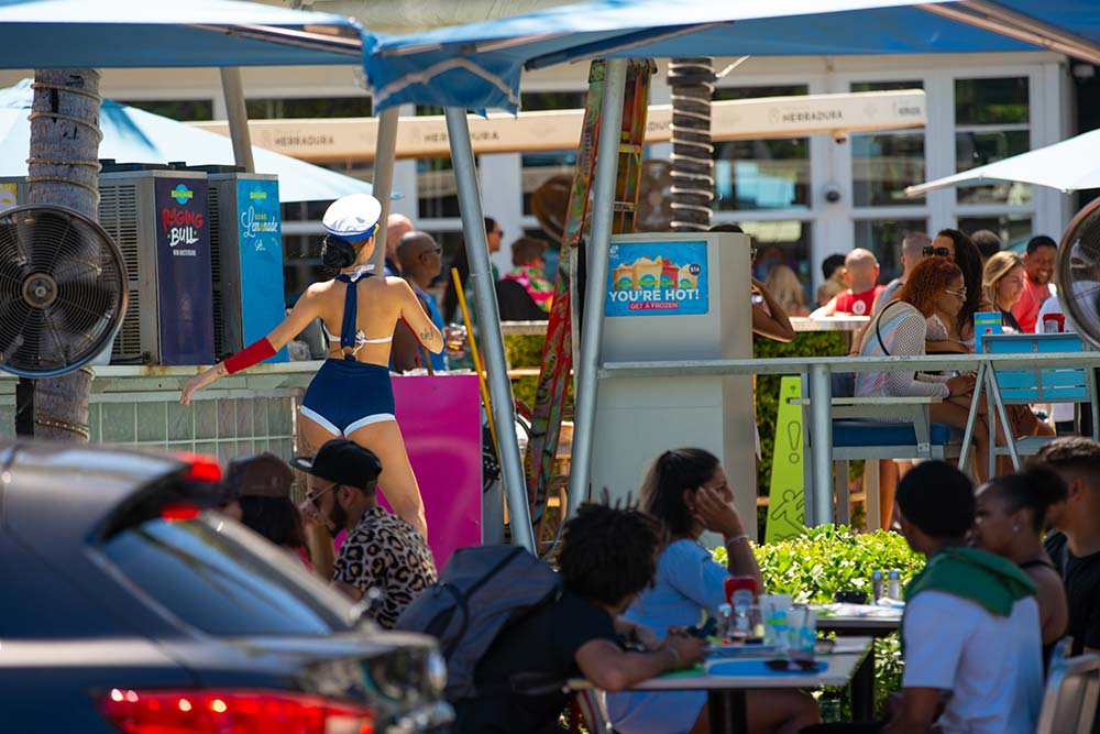 A wide shot of the Clevelander South Beach hotel and bar, with a woman in a sailor costume dancing. Crowds of people are seated at tables in a lively outdoor setting with umbrellas and palm trees.