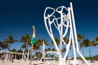 A man in a handstand is balancing on a fitness bar near a large, white, tree-like sculptural fitness installation ("Leopard Tree") on the sandy "Muscle Beach" in Lummus Park, with palm trees and a clear blue sky in the background.