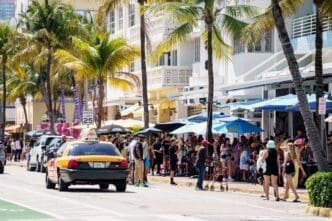 A bustling street scene on Ocean Drive in South Beach, Miami, with a yellow taxi driving by. A crowd of people are seated at outdoor cafes under umbrellas, with palm trees and art deco buildings lining the street on a sunny day.