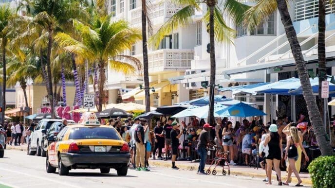 A bustling street scene on Ocean Drive in South Beach, Miami, with a yellow taxi driving by. A crowd of people are seated at outdoor cafes under umbrellas, with palm trees and art deco buildings lining the street on a sunny day.