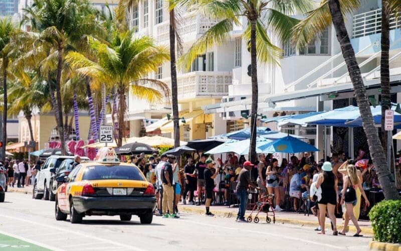 A bustling street scene on Ocean Drive in South Beach, Miami, with a yellow taxi driving by. A crowd of people are seated at outdoor cafes under umbrellas, with palm trees and art deco buildings lining the street on a sunny day.
