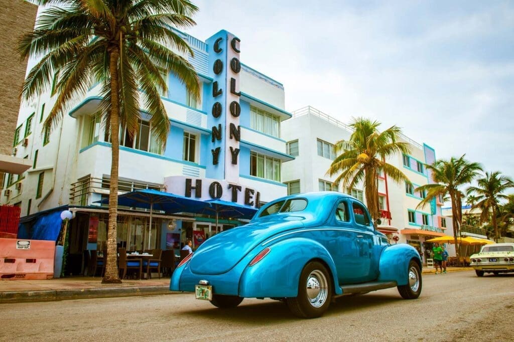 A vintage light blue car drives down Ocean Drive, passing by the iconic Colony Hotel, a white and blue Art Deco building, with palm trees and outdoor seating areas under a cloudy sky.