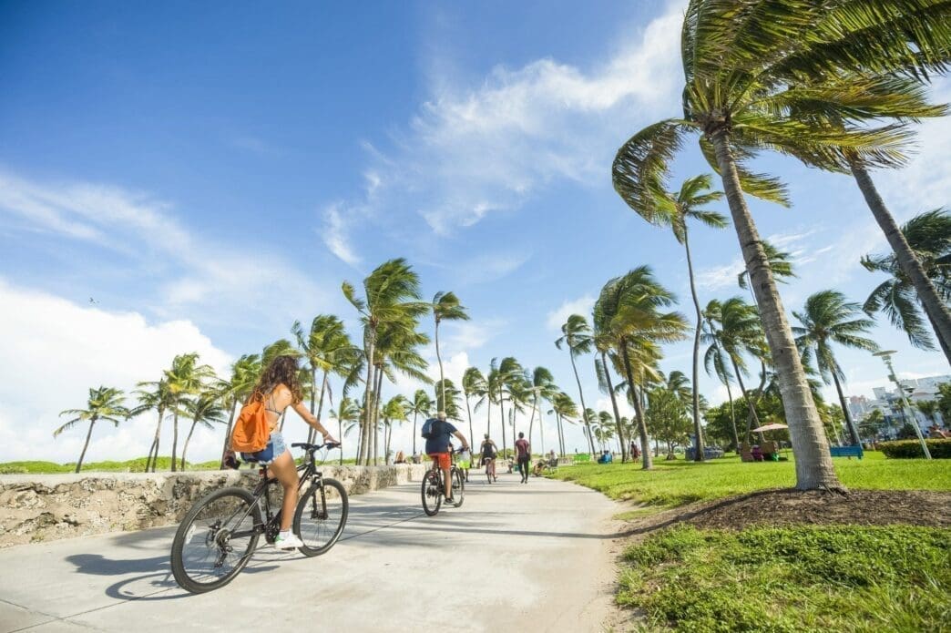 Rear view of a woman inline skating and a man cycling on the Miami Beach Boardwalk, lined with palm trees and buildings.