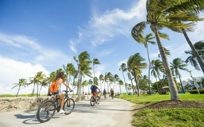 Rear view of a woman inline skating and a man cycling on the Miami Beach Boardwalk, lined with palm trees and buildings.