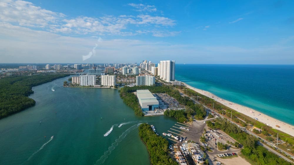An expansive aerial view of Haulover Park in Miami, showing a narrow strip of land with a white sand beach and the turquoise Atlantic Ocean on the right, and the marina and blue waters of Biscayne Bay on the left, with the Sunny Isles skyline in the background.