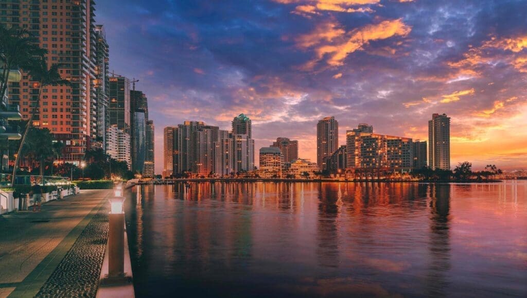 The Miami skyline of Brickell and Brickell Key at sunset, with tall skyscrapers and high-rises beautifully reflected in the calm water of the bay. The sky is a dramatic mix of orange, pink, purple, and blue.