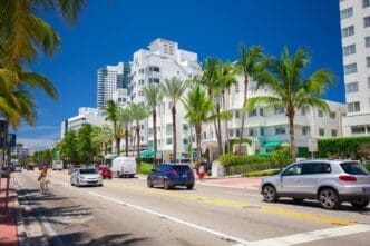 A lively street scene on a sunny day in Miami Beach, with a cyclist and cars on a multi-lane road, framed by tall palm trees and the iconic white Art Deco buildings, including the Hotel Marseilles.