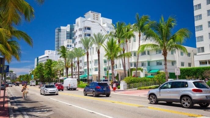 A lively street scene on a sunny day in Miami Beach, with a cyclist and cars on a multi-lane road, framed by tall palm trees and the iconic white Art Deco buildings, including the Hotel Marseilles.
