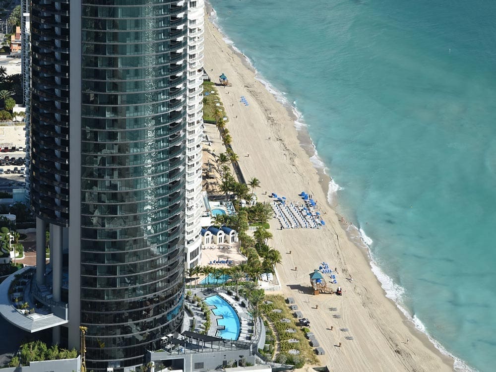 A high-angle aerial view of the Porsche Design Tower in Sunny Isles, Florida, showing the sleek, curved glass facade of the building next to a wide, white sand beach and the turquoise ocean.