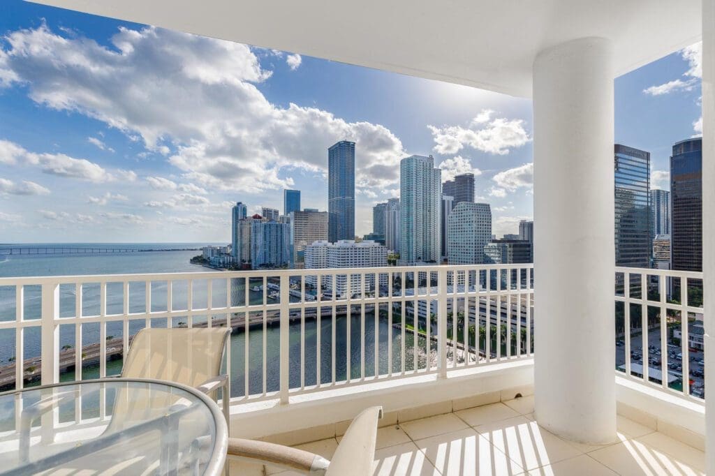 A view of the Miami skyline from a downtown condo balcony, showcasing tall skyscrapers and Biscayne Bay in the background under a partly cloudy sky