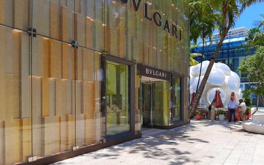 The sleek, modern glass facade of the Bulgari store in the Miami Design District, with palm trees and an interesting bubble-like structure nearby, under a bright sunny sky.