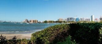 Sarasota Bay with a city skyline and the John Ringling Causeway Bridge, with boats and lush green vegetation in the foreground.
