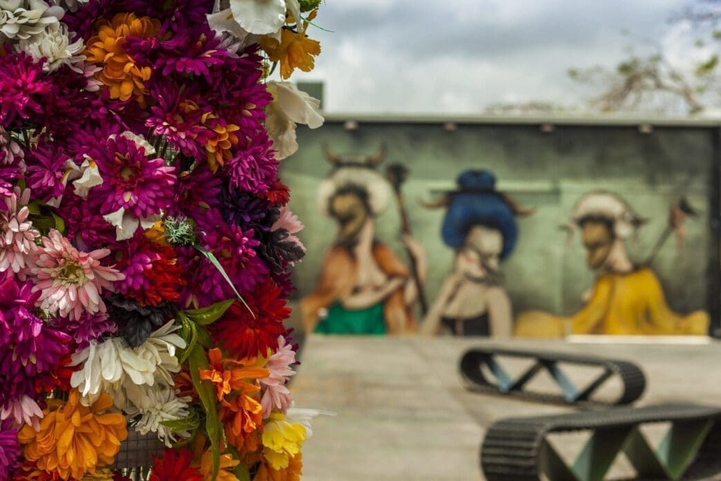 A vibrant floral arrangement in the foreground, with blurred street art murals and a concrete object in the background, typical of Wynwood.