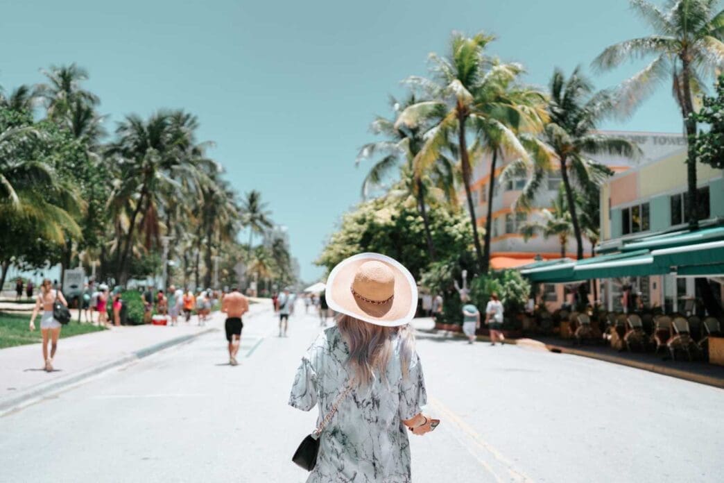 Rear view of a woman in a straw hat strolling down Ocean Drive in Miami Beach, lined with palm trees and Art Deco buildings.