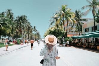 Rear view of a woman in a straw hat strolling down Ocean Drive in Miami Beach, lined with palm trees and Art Deco buildings.