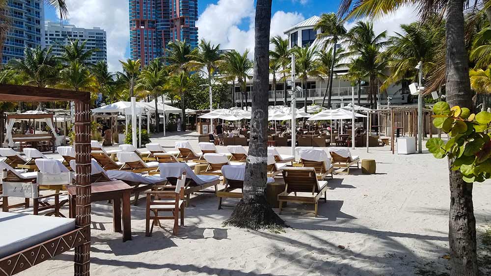 A wide photograph of Nikki Beach in Miami, showing rows of lounge chairs and white cabanas on the sand, with palm trees and modern buildings in the background.