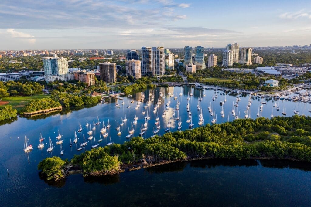 An aerial view of the Coconut Grove neighborhood in Miami, with a large, calm bay filled with numerous sailboats, surrounded by lush green parks and modern high-rise buildings, under a clear sky at sunset.