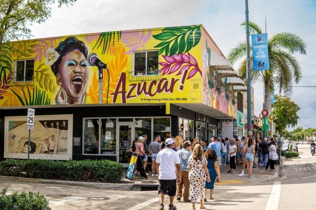 A vibrant street scene in Little Havana, Miami, featuring a colorful mural of Celia Cruz on a building, with people walking by and palm trees.