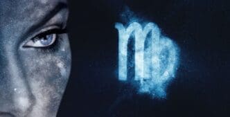 A close-up of a blue-skinned face with a striking blue eye, covered in subtle galaxy-like speckles, looking intensely forward. Beside it, a glowing, light blue, slightly pixelated Virgo zodiac symbol floats against a dark, starry background.