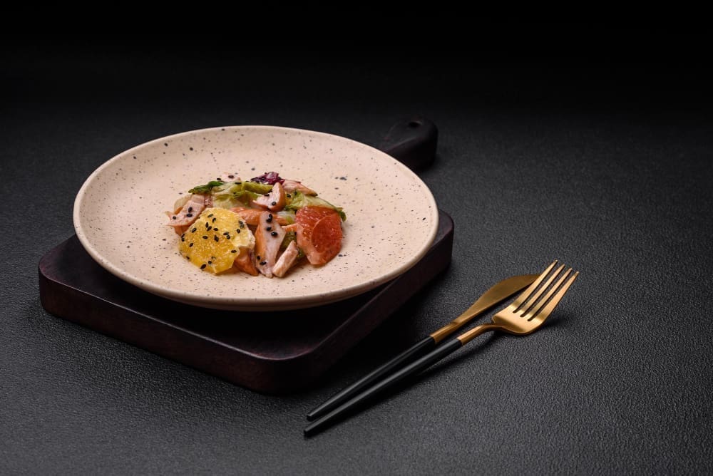 A delectable, fresh salad featuring sliced chicken, grapefruit, lettuce, and sesame, elegantly drizzled with olive oil, served on a ceramic plate