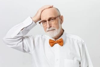 A handsome, bearded retired man in a white shirt and orange bow tie rests his hand on his bald head, looking forlorn after experiencing a stroke