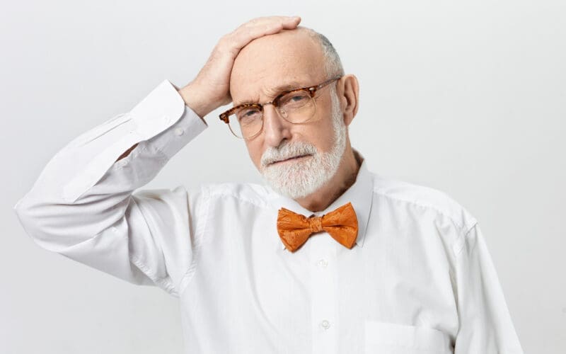 A handsome, bearded retired man in a white shirt and orange bow tie rests his hand on his bald head, looking forlorn after experiencing a stroke