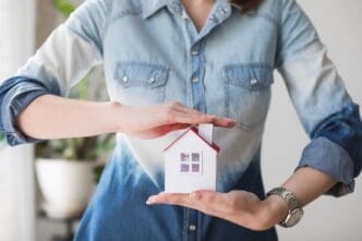 A woman’s midsection shielding a house model, representing the importance of property insurance.