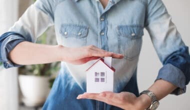 A woman’s midsection shielding a house model, representing the importance of property insurance.