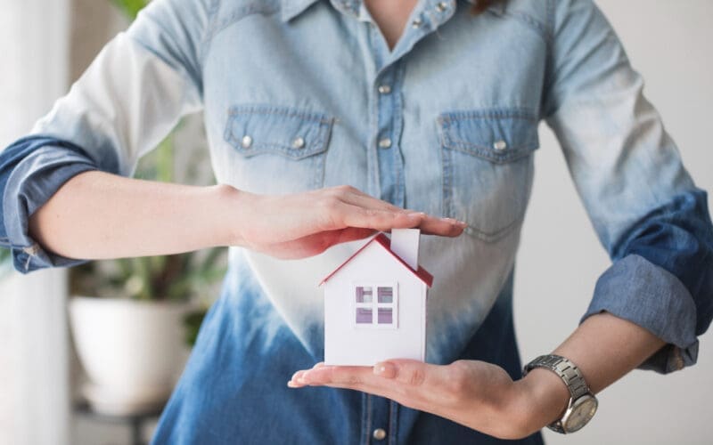 A woman’s midsection shielding a house model, representing the importance of property insurance.