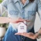 A woman’s midsection shielding a house model, representing the importance of property insurance.