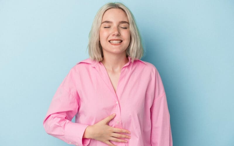 A young Caucasian woman gently touches her tummy while smiling softly, despite experiencing a slight discomfort in her upper abdomen