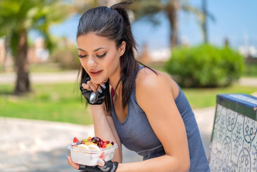 A young, fit woman is holding a bowl of fruit outdoors, preparing to enjoy a healthy snack after her workout