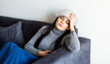 A young woman with migraines is resting on her sofa, a cool, damp towel draped over her forehead.