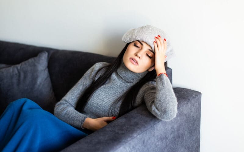 A young woman with migraines is resting on her sofa, a cool, damp towel draped over her forehead.