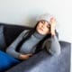 A young woman with migraines is resting on her sofa, a cool, damp towel draped over her forehead.