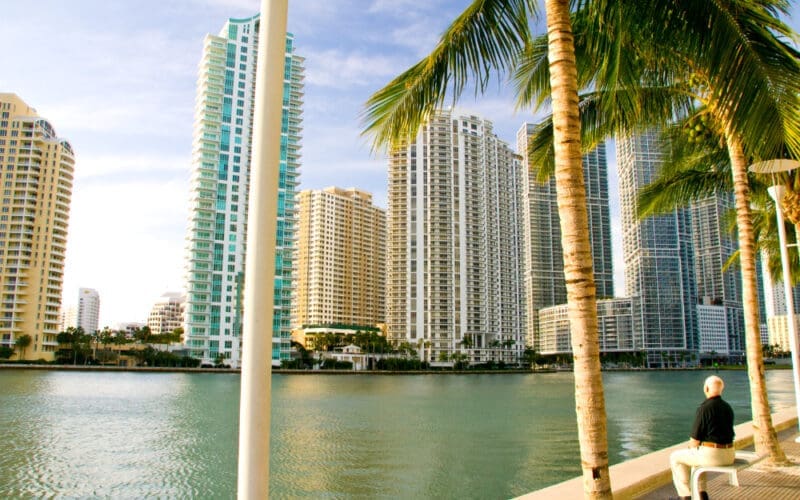 An elderly man relaxes on a bench by Biscayne Bay, taking in the view of the Brickell skyline