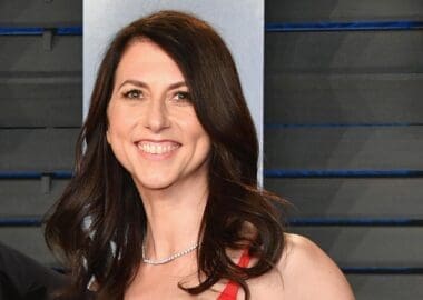 A smiling MacKenzie Scott, with long dark hair and wearing a red top and diamond necklace, looks directly at the camera.