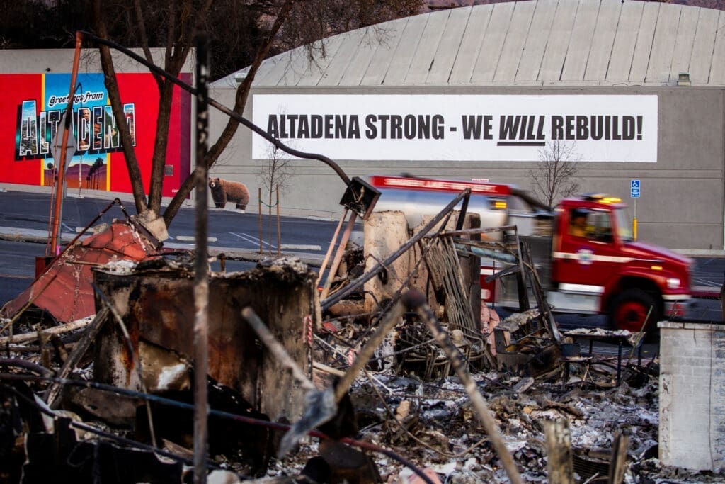 A fire truck passes by banner near structures damaged by Eaton Fire in Altadena