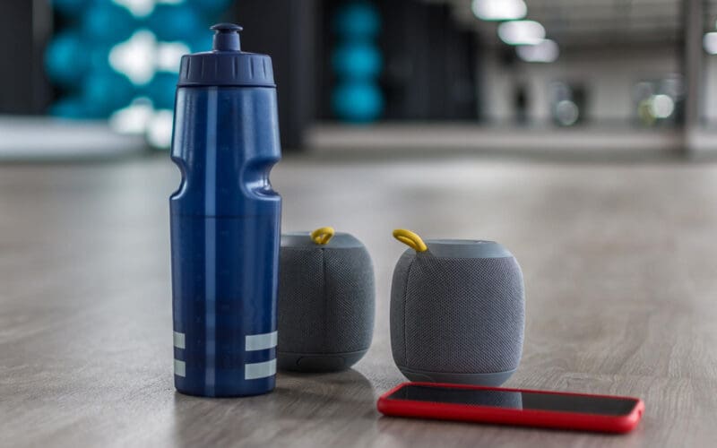 A blue water bottle, portable speakers, and a smartphone resting on the floor of a fitness studio.