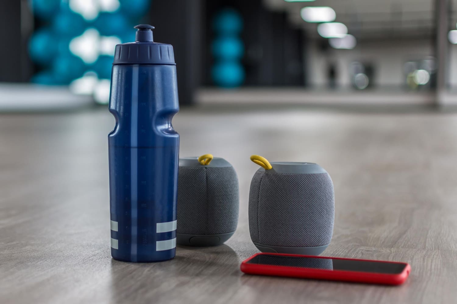 A blue water bottle, portable speakers, and a smartphone resting on the floor of a fitness studio.