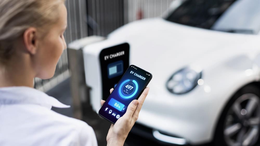 A businesswoman is holding her smartphone, displaying the battery status interface of a smart EV mobile app, while her electric vehicle charges at a station