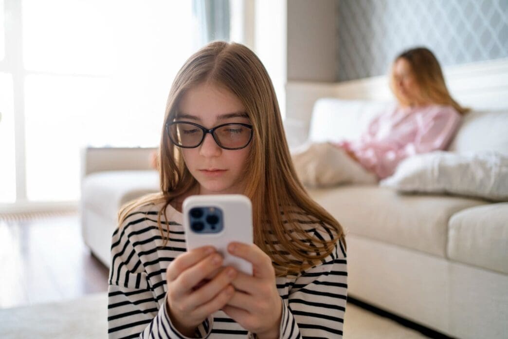 A girl chatting on her cell phone is seen from the front, with her mother seated in the background