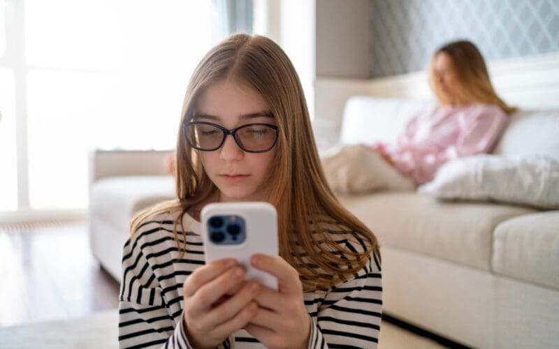 A girl chatting on her cell phone is seen from the front, with her mother seated in the background