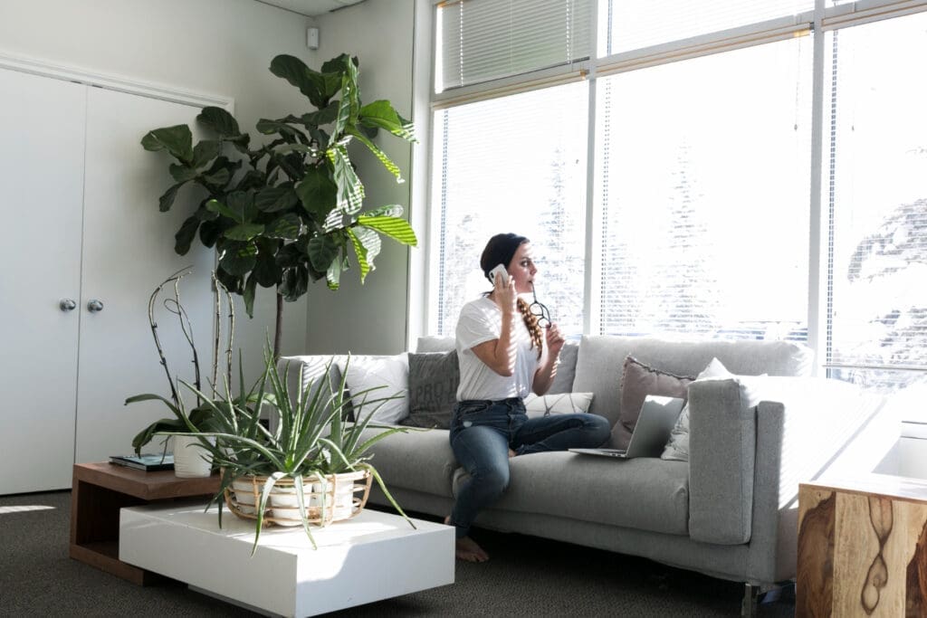 A woman seated on a sofa is talking on the phone, surrounded by potted plants and a tree in the corner of the room