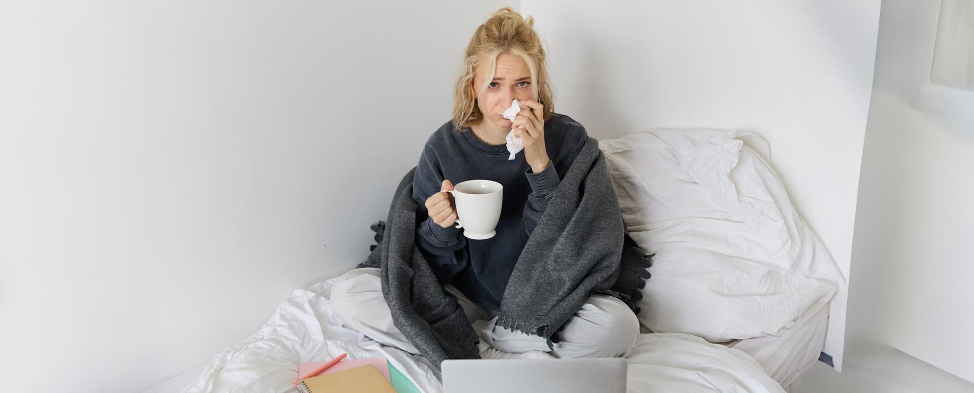 A young woman is at home feeling unwell, sipping tea and passing time in bed